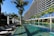 A modern, multi-story hotel with a curved facade overlooking a large swimming pool. Surrounding the pool are several palm trees and neatly arranged lounge chairs with closed umbrellas. The clear blue water of the pool reflects the greenery and the structure of the hotel. The sky is clear and blue.