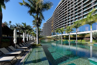 A modern, multi-story hotel with a curved facade overlooking a large swimming pool. Surrounding the pool are several palm trees and neatly arranged lounge chairs with closed umbrellas. The clear blue water of the pool reflects the greenery and the structure of the hotel. The sky is clear and blue.