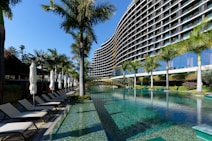 A modern, multi-story hotel with a curved facade overlooking a large swimming pool. Surrounding the pool are several palm trees and neatly arranged lounge chairs with closed umbrellas. The clear blue water of the pool reflects the greenery and the structure of the hotel. The sky is clear and blue.