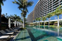 A modern, multi-story hotel with a curved facade overlooking a large swimming pool. Surrounding the pool are several palm trees and neatly arranged lounge chairs with closed umbrellas. The clear blue water of the pool reflects the greenery and the structure of the hotel. The sky is clear and blue.