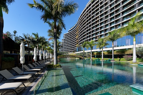 A modern, multi-story hotel with a curved facade overlooking a large swimming pool. Surrounding the pool are several palm trees and neatly arranged lounge chairs with closed umbrellas. The clear blue water of the pool reflects the greenery and the structure of the hotel. The sky is clear and blue.