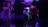 A group of people gathered indoors in a dimly lit environment, likely a party or club setting. The lighting features neon hues of blue and purple, casting colorful shades on the people's clothing. The individuals are engaged in socializing and dancing.