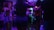 A group of people gathered indoors in a dimly lit environment, likely a party or club setting. The lighting features neon hues of blue and purple, casting colorful shades on the people's clothing. The individuals are engaged in socializing and dancing.