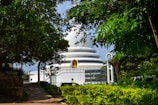 The image depicts a white stupa-like structure surrounded by lush green foliage. There is a sign in the foreground with the text 'Peace Pagoda Rumassala'. The stupa has a tranquil, serene appearance, with a golden statue visible in a niche on its front. A small path leads towards the stupa, and there is a light blue sky in the background.