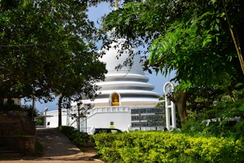 The image depicts a white stupa-like structure surrounded by lush green foliage. There is a sign in the foreground with the text 'Peace Pagoda Rumassala'. The stupa has a tranquil, serene appearance, with a golden statue visible in a niche on its front. A small path leads towards the stupa, and there is a light blue sky in the background.