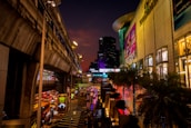 A bustling urban street scene at night with vibrant lights illuminating a shopping area. Elevated train tracks run alongside a large shopping mall adorned with colorful advertisements. Several cars and pedestrians are visible below, adding to the lively atmosphere. Neon signs and illuminated billboards contribute to the city's energetic nightlife.