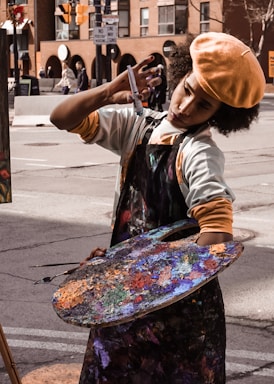An artist is deeply focused while painting on a street, holding a large colorful palette covered in various shades of paint. They are wearing a beret and an apron. The backdrop includes urban buildings and street signs.
