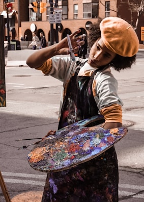 An artist is deeply focused while painting on a street, holding a large colorful palette covered in various shades of paint. They are wearing a beret and an apron. The backdrop includes urban buildings and street signs.