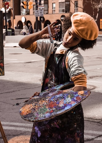 An artist is deeply focused while painting on a street, holding a large colorful palette covered in various shades of paint. They are wearing a beret and an apron. The backdrop includes urban buildings and street signs.