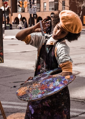 An artist is deeply focused while painting on a street, holding a large colorful palette covered in various shades of paint. They are wearing a beret and an apron. The backdrop includes urban buildings and street signs.