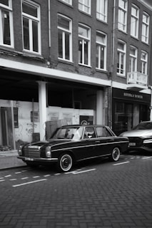 A black vintage car is parked on the side of a cobblestone street, next to a modern vehicle. The setting includes a row of brick buildings with large windows. One of the storefronts has the name 'Bottega Veneta' displayed. Some graffiti is visible on the glass windows of one building.