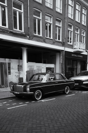 A black vintage car is parked on the side of a cobblestone street, next to a modern vehicle. The setting includes a row of brick buildings with large windows. One of the storefronts has the name 'Bottega Veneta' displayed. Some graffiti is visible on the glass windows of one building.