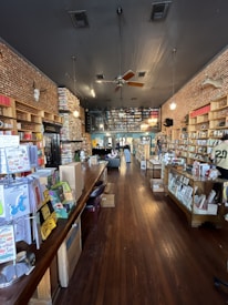 A cozy bookstore interior with wooden floors and walls lined with shelves filled with books. The ceiling has exposed brickwork and lighting fixtures hanging down. A large wooden counter on the left side is covered with various items, and a few people are inside, browsing and relaxing.