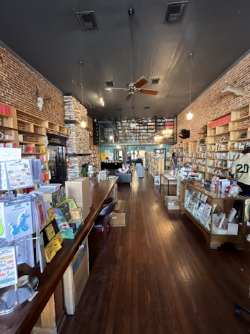 A cozy bookstore interior with wooden floors and walls lined with shelves filled with books. The ceiling has exposed brickwork and lighting fixtures hanging down. A large wooden counter on the left side is covered with various items, and a few people are inside, browsing and relaxing.