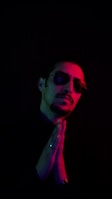 A person wearing sunglasses appears with hands clasped together as if in prayer or contemplation. The image is predominantly dark with dramatic lighting, highlighting the face in shades of red and blue.