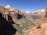 A vast canyon landscape with sheer sandstone cliffs and a clear blue sky. The valley is dotted with green vegetation, and a winding road is visible far below. The rock formations display a range of earthy colors, from deep reds to lighter beige tones.