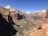A vast canyon landscape with sheer sandstone cliffs and a clear blue sky. The valley is dotted with green vegetation, and a winding road is visible far below. The rock formations display a range of earthy colors, from deep reds to lighter beige tones.
