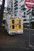 A colorful food truck is parked on a street near high-rise buildings. The back of the truck is painted with bright yellow and displays cartoon images of breakfast items, tacos, fruits, and burritos. Nearby are street signs including 'Do Not Enter' and 'One Way', and the scene is bordered by trees and parked cars.