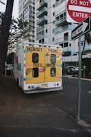 A colorful food truck is parked on a street near high-rise buildings. The back of the truck is painted with bright yellow and displays cartoon images of breakfast items, tacos, fruits, and burritos. Nearby are street signs including 'Do Not Enter' and 'One Way', and the scene is bordered by trees and parked cars.