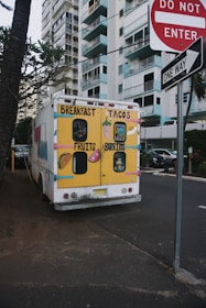 A colorful food truck is parked on a street near high-rise buildings. The back of the truck is painted with bright yellow and displays cartoon images of breakfast items, tacos, fruits, and burritos. Nearby are street signs including 'Do Not Enter' and 'One Way', and the scene is bordered by trees and parked cars.