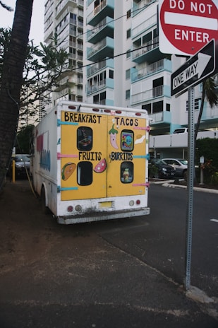 A colorful food truck is parked on a street near high-rise buildings. The back of the truck is painted with bright yellow and displays cartoon images of breakfast items, tacos, fruits, and burritos. Nearby are street signs including 'Do Not Enter' and 'One Way', and the scene is bordered by trees and parked cars.