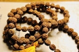 A string of prayer beads made from small, textured brown seeds, intricately arranged in a circular fashion. A yellow tassel is attached at one end, adding a vibrant touch to the earthy tones.