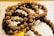A string of prayer beads made from small, textured brown seeds, intricately arranged in a circular fashion. A yellow tassel is attached at one end, adding a vibrant touch to the earthy tones.