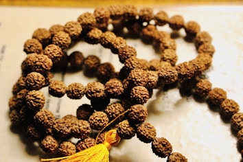 A string of prayer beads made from small, textured brown seeds, intricately arranged in a circular fashion. A yellow tassel is attached at one end, adding a vibrant touch to the earthy tones.