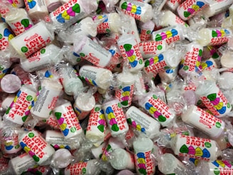 A large pile of small cylindrical candy packets wrapped in transparent plastic. Each packet features colorful heart-themed designs and the words 'Love Hearts' in bold red letters. The candies inside are round and come in various pastel colors.