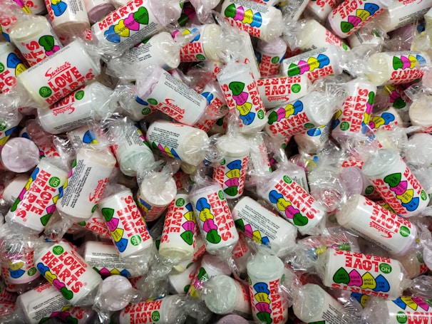 A large pile of small cylindrical candy packets wrapped in transparent plastic. Each packet features colorful heart-themed designs and the words 'Love Hearts' in bold red letters. The candies inside are round and come in various pastel colors.