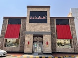 A building exterior with stone-like walls is visible, featuring two large red awnings on either side of a central entrance. Above the entrance is a black square sign with Arabic text in red. Reflective windows on either side show street reflections, while a yellow and black curb lines the street in front of the building.