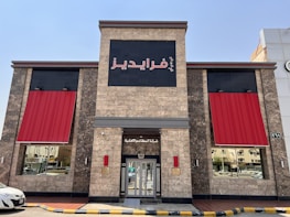A building exterior with stone-like walls is visible, featuring two large red awnings on either side of a central entrance. Above the entrance is a black square sign with Arabic text in red. Reflective windows on either side show street reflections, while a yellow and black curb lines the street in front of the building.