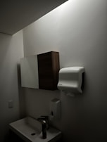 A dimly lit bathroom features a small sink with a tall bottle on it. Above, there is a wall-mounted cabinet with both wooden and mirrored surfaces. A paper towel dispenser is mounted on the wall nearby, along with a soap dispenser below it. The walls and ceiling are light-colored.