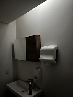 A dimly lit bathroom features a small sink with a tall bottle on it. Above, there is a wall-mounted cabinet with both wooden and mirrored surfaces. A paper towel dispenser is mounted on the wall nearby, along with a soap dispenser below it. The walls and ceiling are light-colored.