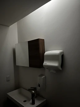 A dimly lit bathroom features a small sink with a tall bottle on it. Above, there is a wall-mounted cabinet with both wooden and mirrored surfaces. A paper towel dispenser is mounted on the wall nearby, along with a soap dispenser below it. The walls and ceiling are light-colored.