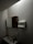 A dimly lit bathroom features a small sink with a tall bottle on it. Above, there is a wall-mounted cabinet with both wooden and mirrored surfaces. A paper towel dispenser is mounted on the wall nearby, along with a soap dispenser below it. The walls and ceiling are light-colored.
