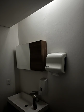 A dimly lit bathroom features a small sink with a tall bottle on it. Above, there is a wall-mounted cabinet with both wooden and mirrored surfaces. A paper towel dispenser is mounted on the wall nearby, along with a soap dispenser below it. The walls and ceiling are light-colored.
