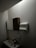 A dimly lit bathroom features a small sink with a tall bottle on it. Above, there is a wall-mounted cabinet with both wooden and mirrored surfaces. A paper towel dispenser is mounted on the wall nearby, along with a soap dispenser below it. The walls and ceiling are light-colored.