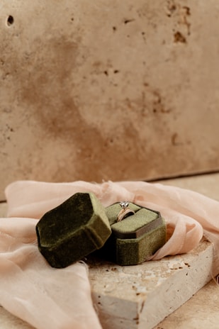 An elegant engagement ring with a heart-shaped diamond sits in an open, green velvet ring box. The box rests on a piece of stone and is surrounded by a soft, blush pink fabric. The background is a textured, neutral-colored surface.