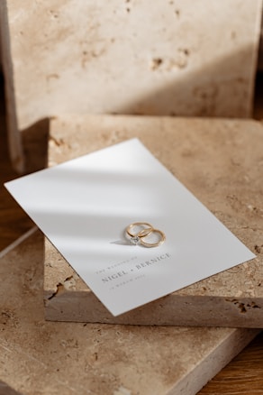 Two gold rings rest on a white card with elegant text indicating a wedding invitation, placed on textured stone slabs.