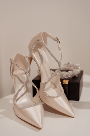 Elegant high-heeled shoes with a satin-like finish and sparkling straps, positioned on a reflective surface next to a small black box containing a crystal accessory.