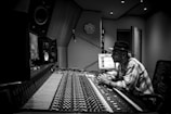 A person wearing a hat is sitting in a music studio, closely observing a computer screen and surrounded by audio equipment, including large speakers and a mixing console. The room has a modern design with acoustic panels on the walls.