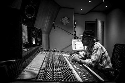 A person wearing a hat is sitting in a music studio, closely observing a computer screen and surrounded by audio equipment, including large speakers and a mixing console. The room has a modern design with acoustic panels on the walls.