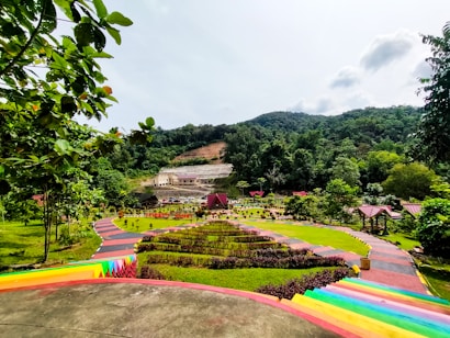 A scenic park landscape with colorful pathways, lush greenery, and hills in the background. The image features vibrant plants and well-maintained gardens, with a small building situated against a backdrop of forested mountains.