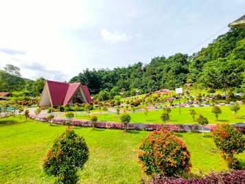 A picturesque landscape featuring a triangular, A-frame house with a red roof in the center. The foreground showcases a lush green lawn with neatly trimmed shrubs and bushes, arranged in symmetrical patterns. The background consists of a dense collection of trees and a hilly area. The sky is bright and slightly cloudy, contributing to the overall serenity of the scene.