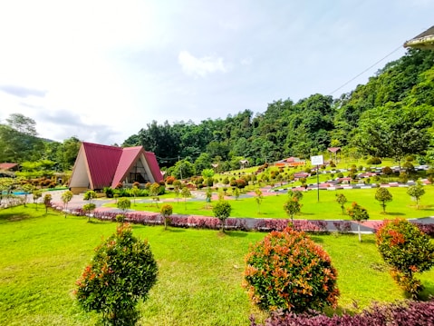 A picturesque landscape featuring a triangular, A-frame house with a red roof in the center. The foreground showcases a lush green lawn with neatly trimmed shrubs and bushes, arranged in symmetrical patterns. The background consists of a dense collection of trees and a hilly area. The sky is bright and slightly cloudy, contributing to the overall serenity of the scene.