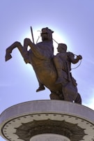 A bronze statue of a warrior on a rearing horse, holding a sword with one hand. The statue is situated on a cylindrical pedestal decorated with intricate patterns. The sky is clear, and the sun creates a halo effect around the statue.