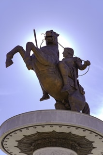 A bronze statue of a warrior on a rearing horse, holding a sword with one hand. The statue is situated on a cylindrical pedestal decorated with intricate patterns. The sky is clear, and the sun creates a halo effect around the statue.