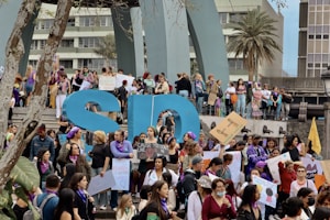 A large group of people, predominantly women, are gathered for a protest or rally. They are holding signs with various messages, many of which focus on women's rights and empowerment. Some participants are wearing purple, a color often associated with feminist movements. The atmosphere is lively and engaged, with a sense of solidarity among the crowd.