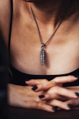 A person wearing a metallic pendant on a beaded chain around their neck. The pendant is elongated and textured, resembling a series of stacked segments. The individual's hands are crossed in front, with fingers interlaced. The lighting creates a moody, intimate atmosphere, highlighting the textures and contours of the skin and jewelry.